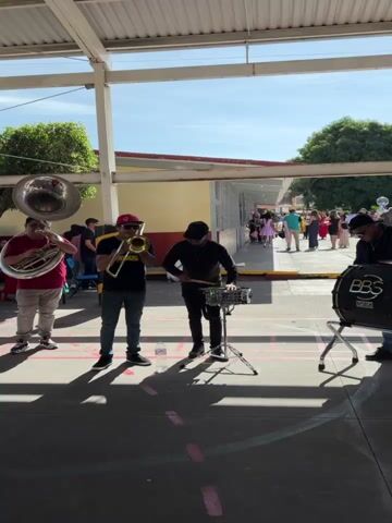 A group of four people are playing instruments outdoors under a covered walkway. The person on the far left is playing a large brass instrument, possibly a tuba. Next to them, a person in a black t-shirt with yellow writing and a red hat is playing a trumpet. In the center, a person wearing a black shirt and pants is playing a snare drum. On the far right, a person in a black shirt and pants is playing a large bass drum. In the background, other people are visible walking in a hallway or open area of a building, some are wearing colorful clothing. The ground is paved and has markings on it, possibly from a basketball court. The sky is visible above the walkway.