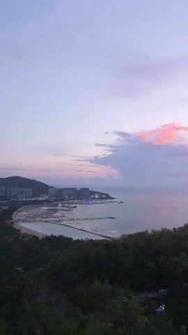 This video captures a serene coastal landscape at dusk. The scene is dominated by a vast expanse of ocean meeting a sky painted with the soft hues of sunset, transitioning from a light lavender to hints of pink and orange. Wispy clouds drift across the horizon, some catching the last rays of sunlight.

In the foreground, a lush, green hillside slopes down towards the water, dotted with trees and foliage that create a sense of natural beauty and tranquility. The hillside appears to be a vantage point, offering a panoramic view of the scene below.

Along the coastline, a city or town is visible, characterized by buildings and structures that reflect the fading light. Notably, three prominent, cylindrical buildings stand out, illuminated with bright red and pink lights, creating a striking contrast with the natural surroundings. Several boats and ships are scattered across the water, suggesting maritime activity. A long pier or breakwater extends into the sea, further defining the shoreline.

The time of day is clearly indicated by the soft, diffused light and the colors in the sky, signifying the approach of evening. The weather appears to be clear with some cloud cover. The presence of a developed coastline with illuminated buildings suggests a populated and developed area, likely a city or town within the Jiyang District of China, given the location context. The overall mood evoked is one of peacefulness and natural splendor, with a touch of urban vibrancy introduced by the illuminated buildings. There is no visible text within the video.