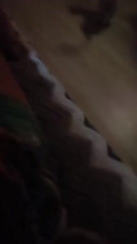 In a dimly lit room with wood flooring, an orange cat is chasing a red laser pointer dot. The cat moves quickly across the floor, its tail raised. In the background, there is a small wooden table and a dark garment hanging on a rack. The camera angle is low, capturing the cat's movements and the red dot on the floor.
