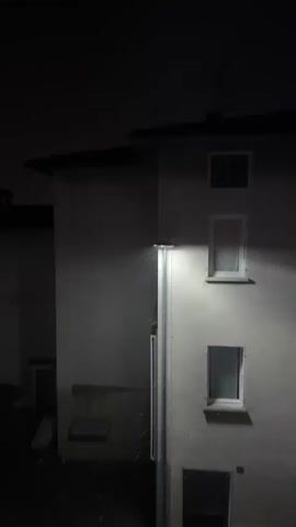 The video shows a building at night with a streetlight illuminating the wall and some falling precipitation, possibly rain or snow. There are several windows visible on the building's facade, some with curtains. Shadows are cast by the streetlight onto the building and the dark sky above, creating streaks of light. The precipitation appears as small white dots against the illuminated background. The overall scene is dark and atmospheric, with the streetlight being the primary source of light.