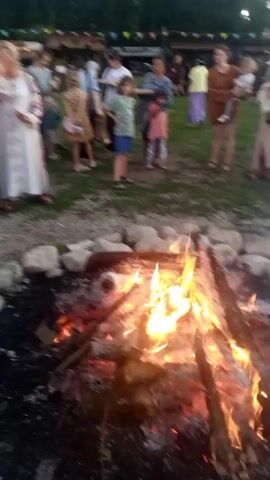 The video shows a gathering of people outdoors, likely a festival or celebration. In the foreground, a bonfire crackles within a ring of stones. Behind the bonfire, a group of people are visible, some holding what appears to be a drum. The individuals are dressed in casual and traditional clothing. The environment suggests a rural or natural setting with trees and hills in the background. The lighting indicates it is late afternoon or early evening. The video is shaky and slightly out of focus, suggesting it was filmed handheld.