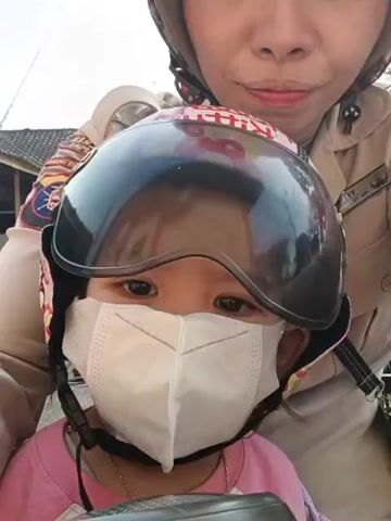 A close-up, selfie-style video shows a woman and a young child on a motorcycle. The woman, in the background, is wearing a light brown uniform shirt with patches on the sleeves and collar. She has dark hair and is smiling. The child, in the foreground, is wearing a pink shirt and a white face mask. The child's helmet is pink with a Hello Kitty design and has a clear visor. The child is looking forward. The background appears to be an outdoor environment with some buildings and vegetation visible. The video is handheld and slightly shaky.