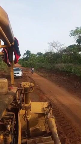 The video shows a wide-angle view from the perspective of yellow heavy machinery, likely a grader or bulldozer, positioned on a reddish-brown dirt road. In the immediate foreground, part of the machinery's engine or hydraulic system is visible, along with some red and black fabric hanging.

Further down the unpaved road, a silver Toyota pickup truck (possibly a Tacoma) is parked on the left side. Its license plate, "MAG-679J", is clearly legible. Two individuals on motorcycles are seen riding along the road in the distance, heading away from the camera's viewpoint. One motorcycle appears to be passing the other.

The road is flanked on both sides by dense green vegetation and trees, including several palm trees, suggesting a rural or semi-rural setting. The sky is bright, indicating daytime. The ground is rugged and uneven, characteristic of an unpaved road in a natural environment. The overall scene depicts activity on a dirt road in what appears to be Otunade, Nigeria, as indicated by the provided context.