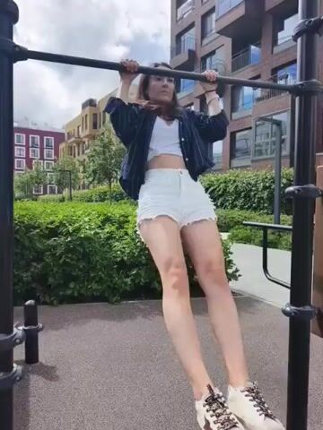 A young woman is performing pull-ups on an outdoor exercise bar. She is wearing white shorts, a white crop top, and a striped jacket. She is using an overhand grip on the bar, with her feet hanging down. The background shows a green hedge and modern apartment buildings.
