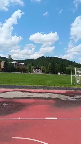 The video displays an outdoor sports complex under a bright blue sky with scattered white clouds. The foreground features a red multi-sport court, visibly marked with wet patches and several strips of light-colored sand or dirt. On the left side of this court, a person wearing a white shirt and dark pants stands, facing towards a distant green grass field. The camera pans to focus on a basketball hoop with a clear backboard, an orange rim, and a white net. The view then shifts across the green field, revealing stadium bleachers, a small white building, and multiple individuals engaged in activities on the field, some appearing to play with a ball. The video concludes with a wider perspective of the red court, showcasing its sandy strips and another basketball hoop positioned further away. Hills covered with trees are visible in the background throughout the scene.
