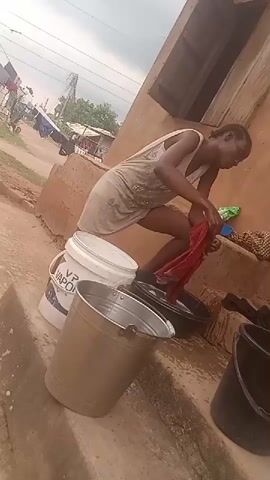 A woman is washing clothes outside a building. She is squatting next to a black basin full of water and using her hands to scrub a red garment. Several buckets are nearby: a white bucket labeled "VP VAP," a silver bucket, and a black bucket. The building's wall is painted a reddish-brown, and a small window is visible. In the background, there is a road, other buildings, and a power pole. The sky is overcast.