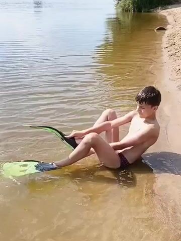 The video shows a young boy sitting on the sandy shore of a lake, with his legs submerged in the shallow water. He is wearing swim trunks and has large green and black flippers on his feet, which he appears to be adjusting. The water is calm with gentle ripples. In the background, the opposite shore is lined with trees, suggesting a natural, wooded environment. The sun is shining brightly, casting a shadow of the boy on the sand, indicating it is likely daytime, possibly late morning or afternoon. The mood is relaxed and casual, suggesting a leisure activity like swimming or preparing to swim. There is no visible text in the video. The location is identified as Nikiforovo, Russia.