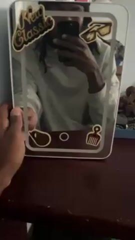 The video shows a person opening a small white refrigerator. The refrigerator is decorated with a mirror on the door with several stickers on it. The inside of the refrigerator contains two bottles of water. The refrigerator sits on top of a brown wooden table. The person’s face is not fully visible, but they are wearing a light-colored long-sleeved shirt. The background is blurry but appears to be a bedroom.