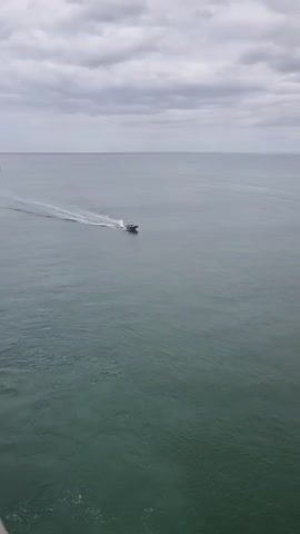 A gray boat creates a white wake as it crosses calm gray-green water under a cloudy gray sky.
