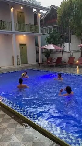 Four individuals are playing in a swimming pool. One person is throwing a soccer ball in the air. The pool is lit with blue lights. There are lounge chairs and umbrellas surrounding the pool. A building with multiple balconies is visible in the background. The weather appears to be overcast or rainy.