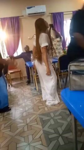 The video depicts a room with blue tables and chairs, where several people are present. Two women are dancing to music; one is wearing a white dress, and the other is wearing a black shirt and blue pants. A man in a navy suit is seated at a table. There are bottles of water, plates with food, and other personal items on the tables. The room has purple curtains, tiled floors, and an air conditioning unit. People are moving around, and the camera shifts its focus between them and the general setting.