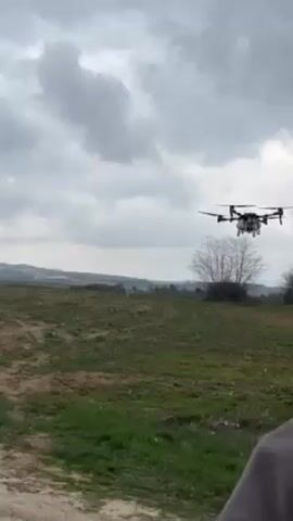 The video shows a drone flying low over a field. The drone is black and white and has multiple propellers. The field is green with some brown patches, and there are some trees and a hill in the background. The sky is cloudy and gray. At the beginning of the video, a person's shoulder and part of their arm are visible at the bottom right corner of the frame.