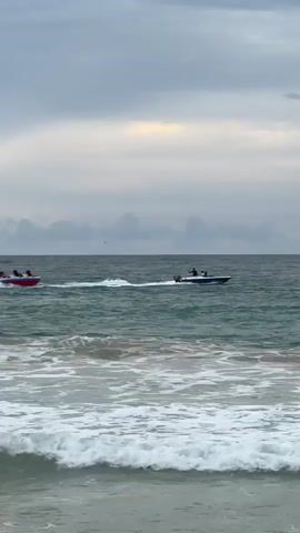 A video shows two boats on the water near the shore. In the foreground, waves are breaking on the beach. In the middle ground, a red and white inflatable tube boat with at least three people on it is being towed by a blue and white speed boat with at least one person visible. The sky is cloudy and gray with hints of orange and yellow in the distance. The water is choppy.