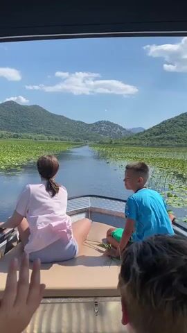 The video opens with a low-angle shot from the back of a boat. Two people, a woman and a young boy, are seated facing forward. The woman, with dark hair pulled back, wears a pink shirt and grey shorts. The boy, with dark hair, is in a blue t-shirt and green shorts. The boat is moving through a body of water covered with lily pads, with a channel of open water in the center. In the background, lush green hills under a bright blue sky with scattered clouds frame the scene. The camera remains relatively still as the boat progresses. A blurred figure's head is visible in the foreground, partially obscuring the view. The boy turns to look at the camera, smiling, while the woman looks ahead. The scene suggests a scenic boat tour on a sunny day.