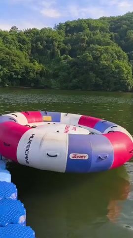 The video shows an inflatable tube labeled "HURRICANE" floating on a body of water. The tube is predominantly white with pink and blue sections. To the left of the tube, a blue floating dock with orange accents is visible. In the background, a densely forested hill slopes down towards the water. The sky is blue with scattered clouds. The camera appears to be on the floating dock, looking towards the inflatable tube and the natural scenery.