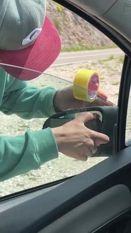 Here is a concise description of the video:

A person wearing a green sweatshirt and a hat tapes a car side mirror to the car window with yellow tape. The person is inside the car and the window is partially down. Outside, there is a paved road and a gravel area next to it. There is a green, rocky hill in the distance. The person's smartphone, encased in a clear case, is visible in the reflection of the side view mirror.