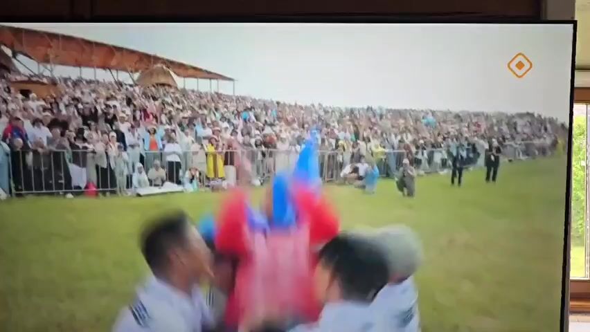 A group of people, some wearing white shirts and hats, are gathered in a grassy field. They are holding up what appear to be large, colorful objects, possibly flags or banners. The objects are red and blue. There are many spectators in the background, seated in bleachers and standing behind a fence. The overall scene suggests a celebratory event or competition.
