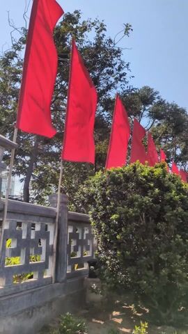 The video shows a row of red flags blowing in the wind against a bright blue sky. The flags are mounted on wooden poles and line a grey concrete fence with a decorative geometric pattern. Behind the fence is a lush green bush and trees. The camera pans slightly to the right, revealing a paved walkway and a large structure in the distance. The day appears sunny and clear.