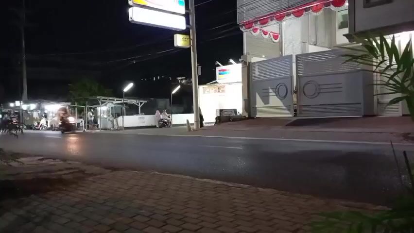 The video shows a street scene at night.  There are several motorcycles on the road, some of which are moving at speed, causing motion blur.  Streetlights illuminate the scene, and there are also lights from various establishments along the side of the road, including food stalls.  A building with a large, illuminated sign is visible on the right side of the frame, along with a large metal gate.  A paved sidewalk is in the foreground.