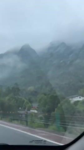 The video shows a view from inside a car driving on a highway.  The road has multiple lanes and the camera is looking forward.  There are other cars on the road, including a large truck in front of the camera car. The environment is cloudy and overcast, with green forested mountains visible in the background. The mountains are partially obscured by low-lying clouds or fog. There are trees lining the side of the road. There is a blue sign visible on the side of the road with writing in Chinese.