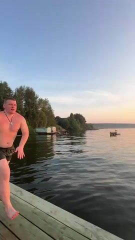 The video shows a person jumping off a wooden pier into a body of water at sunset. The person is wearing dark shorts. The water is calm and reflects the orange and yellow hues of the sunset. A white boat is visible in the distance. The pier is made of wooden planks and appears to be on a lake or a wide river. The sky is clear with a few wispy clouds. In the foreground, part of a green and white checkered blanket and a toy are visible, suggesting someone is filming from the pier. The video captures the action of the jump from two different perspectives, the first from beside the person about to jump and the second from a first-person perspective of someone watching the jump.