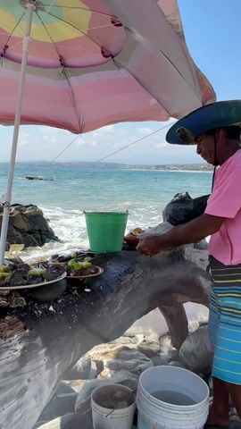 A man in a straw hat, pink shirt, and striped swim trunks stands in front of a stand constructed from driftwood, preparing food under a striped umbrella at a beachside stall. There are plates of food, limes, and buckets of water visible on the stand. The ocean is in the background, with some visible land in the distance. A woman in a black sports bra and white shorts approaches the stall and looks at her phone. The scene is sunny, with puffy clouds in the sky.