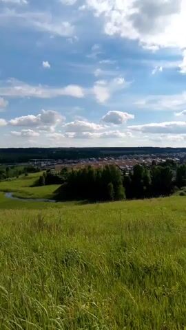 The video shows a green, grassy hill in the foreground. In the mid-ground, there is a winding river and a cluster of trees. In the background, there is a town with houses and a line of dark trees. The sky is blue with white clouds.