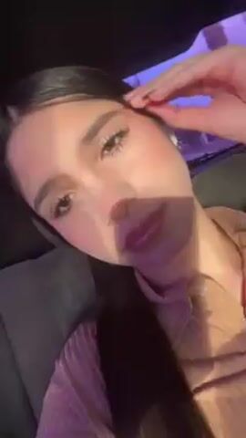 A young woman with long dark hair, fair skin, and wearing lip gloss and an earpiece is in the back seat of a car. She is wearing a light-colored collared shirt. The camera is held close to her face and she looks at it. Outside the car window, there are trees and streetlights.