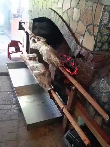 Two whole animals, likely lambs or goats, are skewered on long wooden poles and are being spit-roasted over an open fire inside a stone oven. The animals appear to be wrapped in some sort of casing or stuffing. A metal tray is positioned beneath the roasting animals to catch drippings. The environment appears to be an outdoor or semi-outdoor cooking area with a stone oven and tiled flooring.