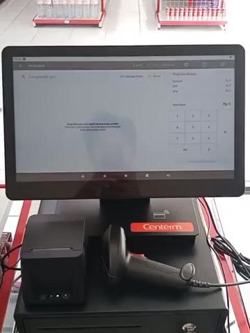 The video shows a point-of-sale system in a retail environment. The system consists of a touchscreen monitor, a barcode scanner, a receipt printer, and a cash drawer. The monitor displays a sales application with fields for searching products, a list of items, and a summary of charges. The screen also shows a message prompting the user to scan a barcode or manually enter a product code. The background of the scene appears to be a store aisle with shelves stocked with various products, including bottles and boxes. The overall impression is that of a functional retail checkout area.