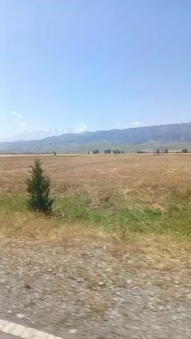 The video captures a moving perspective, likely from a vehicle, traveling along a dirt road. The scenery is predominantly rural, with a large, dry, harvested field occupying the mid-ground. In the distance, rolling hills or mountains are visible under a clear blue sky with scattered clouds. A tall, light-colored pole stands on the right side of the frame. Vegetation, including grass and trees, lines the side of the road and the edge of the field. The camera pans slightly, showing the changing landscape as the journey continues. The overall impression is one of a sunny day in a vast, open, agricultural or semi-arid region.