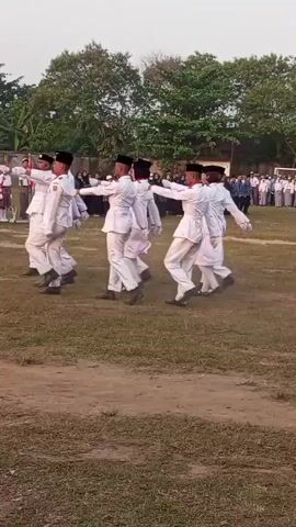 A group of young people in white uniforms with black hats march in formation across a grassy field. They are performing a drill or parade, with synchronized movements. In the background, a larger group of people in dark uniforms watch from a distance. Trees and buildings are visible behind the spectators. The sky is overcast, suggesting it might be morning or late afternoon. The overall scene appears to be an outdoor event, possibly a school ceremony or competition.
