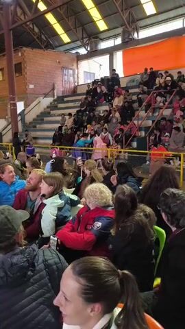 The video shows an indoor event at what appears to be a gymnasium. A seated crowd fills the lower portion of the gymnasium, while the bleachers in the background are also occupied by spectators. There is natural light coming in through windows in the roof structure. People in the crowd are interacting with each other. Someone may be taking photos or video with their phone. The overall atmosphere suggests a community gathering or event.