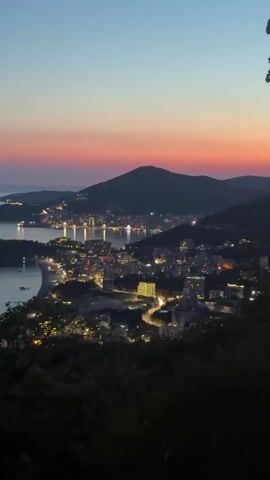 The video shows a panoramic view of a coastal city at dusk. The sky is a gradient of pale blue at the top, transitioning to pink and orange near the horizon. The city lights twinkle below, illuminating the buildings and streets. A dark, forested hill occupies the foreground, framing the scene. The sea is visible to the left, with a small bay and a beach. A few boats are moored in the water. The overall impression is one of serene beauty as day transitions into night.
