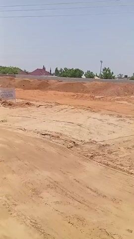 The video pans across a dusty, unfinished construction site under a clear, bright sky. The ground is primarily composed of sand and loose earth, with tire tracks visible from vehicles. In the background, there are piles of sand, gravel, and reddish-brown soil. Several construction materials, including large cylindrical tanks, are scattered around. Some basic structures, appearing to be incomplete buildings or sheds, are also present. Power lines and utility poles are visible overhead. The overall impression is that of a developing area undergoing significant earthworks and infrastructure preparation. No people are clearly visible or engaged in any specific actions.