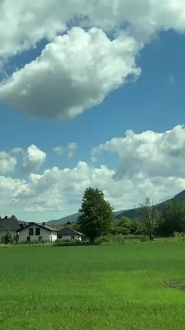 The video shows a view of a rural landscape under a blue sky filled with puffy white clouds. In the foreground is a vibrant green field. Several houses are scattered in the midground, partially obscured by trees. In the distance, gently rolling hills and mountains can be seen.  The camera moves slowly, capturing a tranquil, sunny day in the countryside.