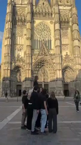 The video shows a group of about 10 young people standing in a circle, in front of a large cathedral. One individual stands on top of the shoulders of the group, and is closest to the cathedral in the frame. The cathedral appears to be constructed from a light beige stone, with significant gothic detail. A person walks across the square in the background. The sky is blue with a few wispy clouds. The individuals in the group are wearing dark shirts, with differing colors and cuts of pants.
