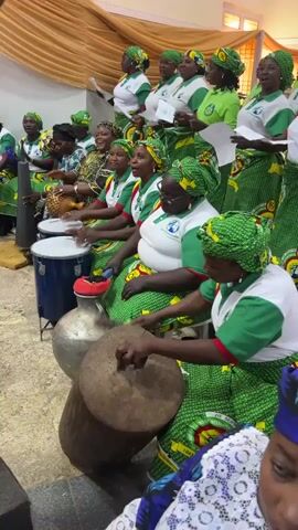A group of women, most wearing matching green and white uniforms and headscarves, are arranged in rows and appear to be singing. Some women are playing percussion instruments, including drums and a shaker. One woman in the foreground is hitting a large wooden drum with a mallet. Another woman to her left is playing a smaller drum with red and black sticks. Several women are holding sheet music. The women are seated on the floor or on low stools. The background appears to be an indoor space with beige walls and curtains.