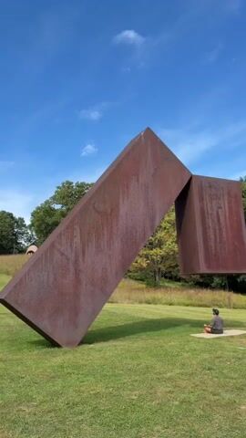 A person sits in a meditative pose on a mat outdoors. They are facing away from the camera, with their legs crossed and hands resting on their knees. They are wearing a dark t-shirt and light-colored shorts. The person is positioned on a grassy field, near a large, rust-colored metal sculpture. The sculpture consists of two large, angular geometric shapes, one of which appears to be suspended in the air. The background shows trees and a clear blue sky with some clouds. The overall scene suggests a peaceful and artistic environment.