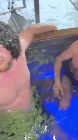 Two men are in a tub of water illuminated with blue lights. The man on the right is wearing black swim trunks and a dark grey beanie. He is holding onto the side of the tub with his right hand. The man on the left has a bushy, black wig on and is holding a vape pen to his mouth. There are some green plants in the tub. The man in the wig gives a thumbs up to the camera. The tub is outdoors, possibly at night.