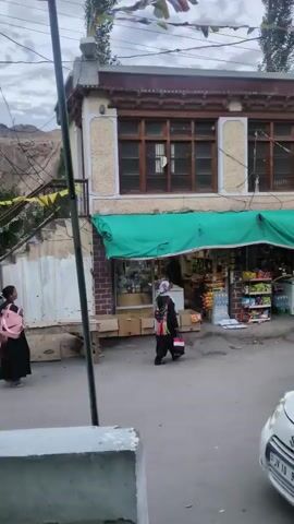 This is a still shot from a video showing a street scene in front of a shop. The shop has a green awning, and shelves stocked with goods are visible inside. Several people are present. On the left, a person in dark clothing with a pink backpack is standing. In the center, a person wearing a headscarf and dark clothing walks towards the shop. To the right, two people, one in dark clothing and the other in a yellow jacket, are standing. A white car with license plate JK 10 9523 is parked in the foreground. Prayer flags are strung across the top of the frame, suggesting a cultural or religious context. The background shows buildings and trees under a cloudy sky.