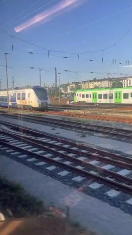 The video shows a view from a train window looking out at a railway yard. In the foreground, multiple sets of train tracks stretch into the distance. Beyond the tracks, several trains are visible.

The first train, on the left side of the frame, is a passenger train with a blue and white color scheme. It appears to be stationary. To its right, another passenger train is visible, predominantly white with blue accents and the text "National Express" on its side. Further down the tracks, several locomotives are lined up. These are mostly white with some colored accents, and one has red and blue sections. In the background, there are buildings and utility poles with overhead electrical wires, common for a train station or yard.

The scene appears to be set during the daytime, with a clear blue sky and bright sunlight casting shadows. The overall atmosphere is one of a working railway environment. There are no people clearly visible in the frame. The camera perspective is from inside another moving train, as indicated by the reflection on the window and the slight motion blur.

The visible text includes "National Express" on one of the trains. Other text is present on the trains but is too small or blurry to read. The location context of Hagen, Germany, suggests this is a German railway facility.
