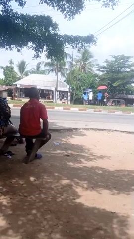 The video shows a scene in what appears to be a tropical or subtropical area, near a road. Two men are sitting on a wooden bench under the shade of a tree. One man is wearing a patterned shirt, and the other is wearing a red shirt. A third man approaches and stands near them, wearing a grey shirt. Across the road, there are people walking, some under colorful umbrellas. In the background, there are buildings and palm trees. A red motorcycle, branded "TIGER," is parked near the bench, beside a small structure. The ground near the bench is sandy. The overall scene suggests a roadside or community gathering place.