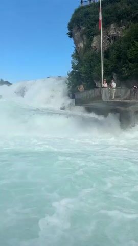 This media file captures a dynamic view of a powerful waterfall, likely the Rhine Falls in Nohl, Switzerland. The scene is dominated by the immense volume of water crashing down, creating a misty spray that fills the foreground with turquoise hues. In the mid-ground, a wooden viewing platform is visible, with several individuals observing the falls from a safe distance. The platform is situated on a rocky outcrop covered in lush green vegetation. A Swiss flag flutters from a flagpole on the cliff face above the platform.

The sky is a clear, vibrant blue with scattered white clouds, suggesting fair weather and likely daytime, given the bright lighting. The water's turbulent movement and the spray indicate a strong flow. The presence of people on the viewing platform and in what appears to be a boat in the foreground suggests a tourist attraction. The overall mood is one of awe and appreciation for the natural spectacle. There is no visible text within the media file.