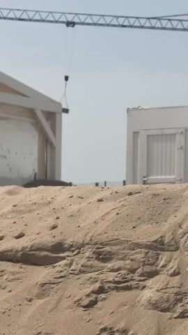 A wide shot shows a sand dune in the foreground, with a building structure and a portable building in the background. A construction crane is visible against a clear sky, with a dark object suspended from its hook. The scene appears to be a beach construction site.