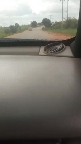 A POV video shows a view from inside a vehicle looking forward through the windshield. The interior of the vehicle, consisting of a gray dashboard and a dark pillar, occupies the foreground. A silver-colored circular object, possibly a hubcap or a decorative car accessory, is situated on the dashboard. The exterior view reveals a paved road with a red car in the distance. The road is flanked by green vegetation and trees under a cloudy sky. The vehicle appears to be in motion, indicated by the slightly blurred scenery.