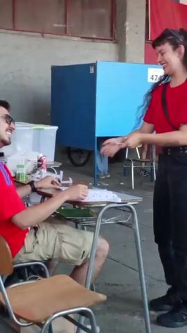 The video shows a man sitting at a desk at a polling place. He is wearing a red and white soccer jersey and khaki shorts. He is examining a stack of papers on the desk in front of him. There are two other people sitting at the table with him, and another person standing behind them. The polling place has blue voting booths and a Chilean flag is visible in the background. A sign with the number 475 is on the wall.