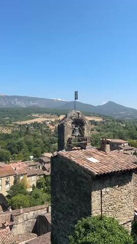 A bell tower stands tall against a clear blue sky. The tower is made of stone and has a bell hanging inside. A flag flies from the top of the tower. In the background, rolling hills and trees create a picturesque landscape. The video is taken from a high vantage point, looking down on a village with red-tiled roofs. The sound of the bell ringing is heard throughout the video.