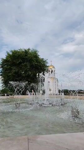 This is a video of a fountain in front of a white building with a golden dome, likely a church, in Kurgan, Russia. The fountain is active, with water spraying upwards and outwards in a dynamic display. Behind the fountain and to the left, a large, lush green tree stands prominently. The building in the background features a cross on top of its dome, reinforcing its religious nature. A few blurry figures can be seen in the distance, possibly people walking near the building. The sky is cloudy, suggesting a diffused light, possibly late morning or afternoon. The scene appears peaceful and serene, with the activity of the fountain providing a sense of gentle movement and life. No visible text is present in the video.