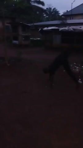 The video shows a dimly lit outdoor scene, likely in Akokwa, Nigeria. The primary subject is a young person, possibly a child or teenager, engaged in playful movement or dancing. They are dressed in dark clothing, including a t-shirt and trousers. The person is seen in various dynamic poses, including jumping and bending, suggesting an energetic activity.

The setting appears to be a rural or semi-urban area. In the background, there are simple structures that resemble houses or makeshift dwellings, some with corrugated metal roofs. Vegetation, including trees, is visible, particularly in the upper portion of the frame. The ground is dark and appears to be made of dirt or earth, with some debris scattered in places.

The time of day is ambiguous due to the low light conditions, but it appears to be either dusk or dawn, or possibly a heavily overcast day. The lighting is very poor, creating a shadowy and somewhat indistinct image. The weather is not explicitly clear, but the overcast conditions might suggest a possibility of rain or recent rain.

There are no discernible text elements within the video. The overall mood is one of youthful energy and playfulness, contrasted with the muted and somewhat somber visual quality of the footage due to the low light. The focus of the video is clearly on the individual's activity and their interaction with the space.